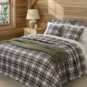 Woody green and red plaid quilt on a bed with matching plaid pillows and a green fringed throw in a wood-panelled bedroom.